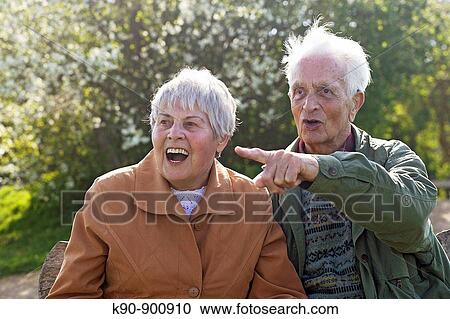 Stock Image - Elderly couple sitting on a bench, looking astonished with open mouth while the senior man pointing with his finger, Island of Amrum, Northfrisian, Schleswig_Holstein, Northern Germany, Europe. Fotosearch