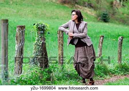Picture - Elegant woman standing up on a forest road close to a fence. Fotosearch