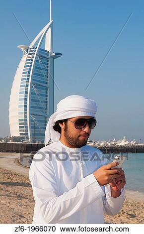 Stock Image - Emirate arab man with cell phone texting in front of Worlds only 7 star hotel in Dubai UAE called the Burj Al Arab with luxury and water at beach in United Arab Emirates.. Fotosearch