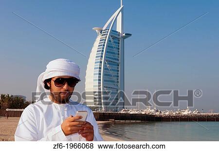 Emirate arab man with cell phone texting in front of Worlds only 7 star hotel in Dubai UAE called the Burj Al Arab with luxury and water at beach in United Arab Emirates. View Large Photo Image Stock Photo - Emirate arab man with cell phone texting in front of Worlds only 7 star hotel in Dubai UAE called the Burj Al Arab with luxury and water at beach in United Arab Emirates.. Fotosearch