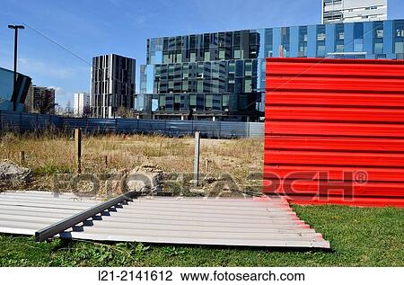 Stock Image - Fallen red aluminium wall, green grass, wasteland, fence, office buildings, blue sky. Pla? a Europa, Plaza Europa, District VII, Gran Via, Hospitalet de Llobregat, Barcelona province, Catalonia, Spain.. Fotosearch