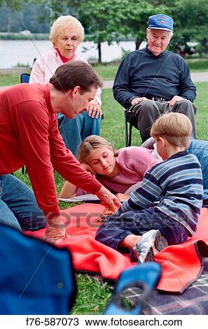 Stock Image - Family, including father and grandparents, camping, on lawn chairs, playing checkers.. Fotosearch