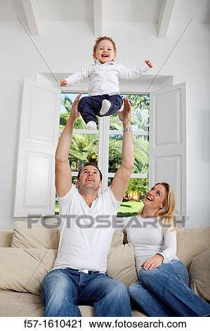 Stock Image - Family on their sofa throwing one year old baby in the air. Fotosearch