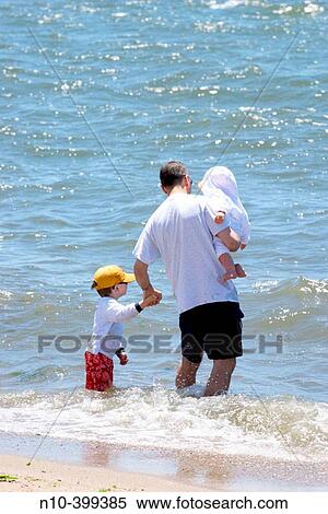 Stock Photography - Father with young sons, one held in his arm and the other holding his hand, standing in the surf at Hammonasset Beach in Madison, Connecticut.. Fotosearch