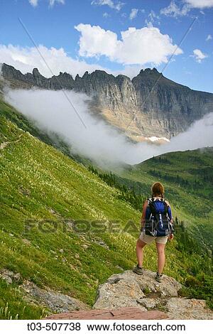 Stock Photo - Female hiker pausing on rock outcrop to take in view of the Garden Wall as seen from the Highline Trail in Glacier National Park, Montana, USA. Fotosearch