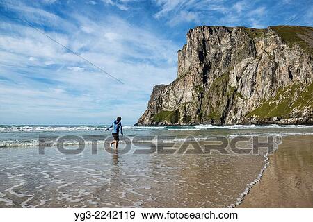 Stock Photo - Female hiker walks in sea below Ryten at Kvalvika beach, Moskenesoy, Lofoten Islands, Norway.. Fotosearch