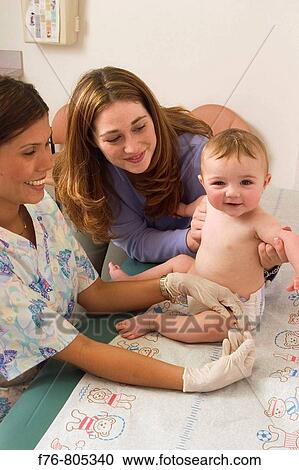 Stock Image - Female pediatric nurse giving injection into thigh of infant, with mother, comforting and looking on.. Fotosearch