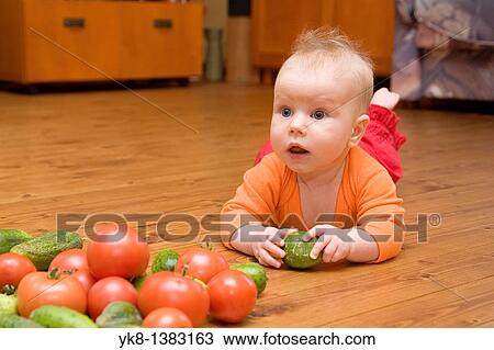 Stock Image - Five month old infant girl lying down on wooden floor, looking and playing with vegetables. Fotosearch