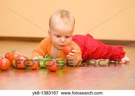 Stock Photo - Five month old infant girl lying down on wooden floor, looking and playing with vegetables. Fotosearch