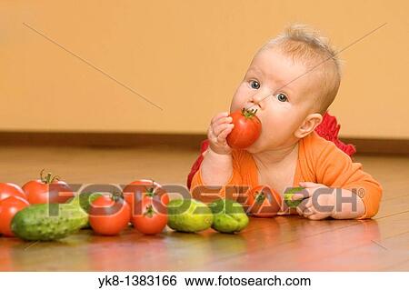 Stock Photograph - Five month old infant girl lying down on wooden floor eating tomato. Fotosearch