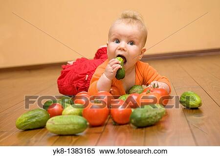 Stock Photography - Five month old infant girl lying down on wooden floor, looking and playing with vegetables. Fotosearch