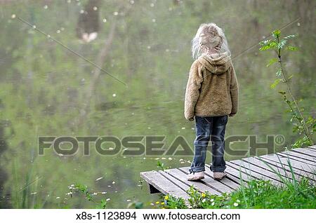 Picture - Four year old girl standing on her own by the side of a pond. Fotosearch