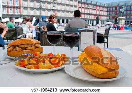 Stock Image - Fried squids sandwich, patatas bravas and Spanish omelet sandwich with two glasses of red wine in a terrace. Main Square, Madrid, Spain.. Fotosearch