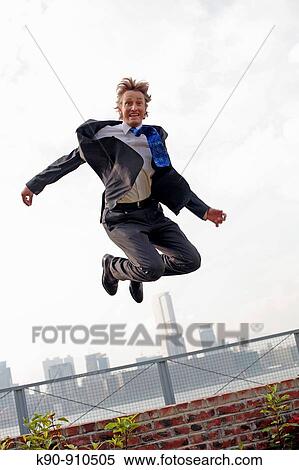 Stock Photography - Full body photograph of young, caucasian business man in business suit, jumping over a fence, with highrise office buildings in the background, Hong Kong, China, Asia. Fotosearch