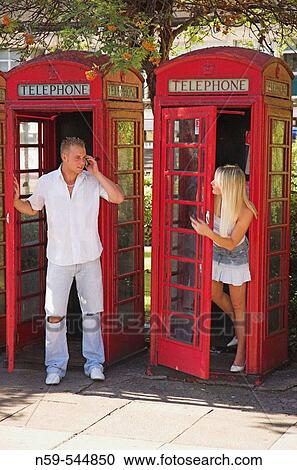 Stock Image - Fun young couple using mobile phones around British public phone boxes.. Fotosearch