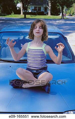Stock Image - Girl about 9 years old in tank top and shorts sitting cross-legged on hood of light blue car with eyes closed, arms out to sides with elbows bent, in meditation posture. Fotosearch