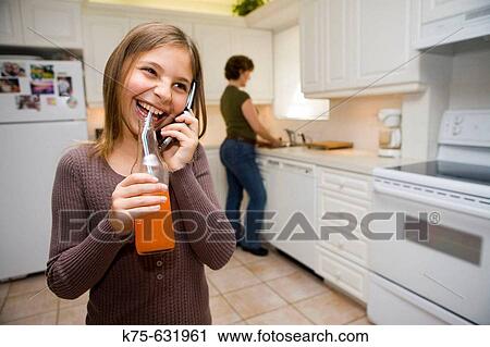 Stock Image - Girl in kitchen with mom in background.. Fotosearch