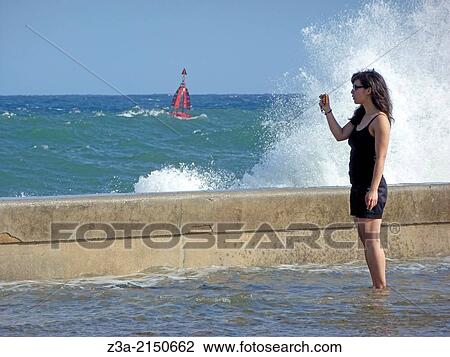 Stock Image - Girl making pictures of the high waves in stormy weather, located on the Malec? n in Havana, Cuba.. Fotosearch