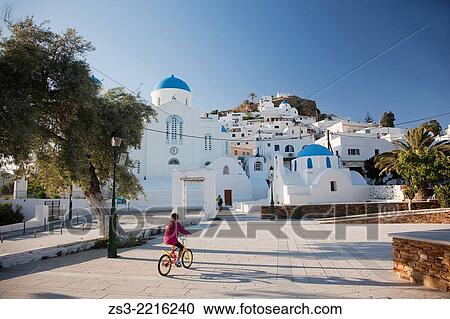 Stock Image - Girl riding a bike near a typical Cyclades church, a blue domed white Byzantine Greek Orthodox Chapel of Panaghia Gremiotissa in Chora, Ios, Cyclades Islands, Greek Islands, Greece, Europe.. Fotosearch