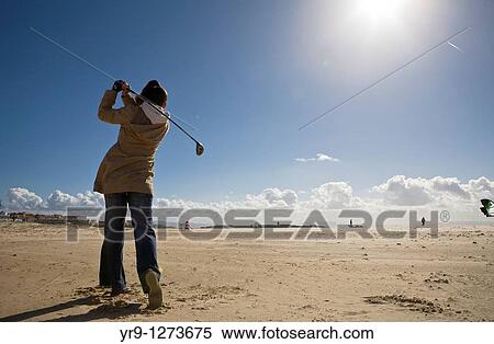 Stock Photography - Golf training doesn? t have to be on a green. An uncrowded beach in winter is actually a perfect place to do it. It even has its special appeal for its weirdness, given the presence -in this case on Tarifa beach, Spain- of kiteboarders in the background. Fotosearch