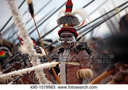Stock Image - Half body image of local Papuan man, looking at the camera, with traditional body painting, impressive headdress and wild boar teeth put through his nose, carrying his bow and arrow, Baliem Valley festival, Jayawijaya region. Fotosearch