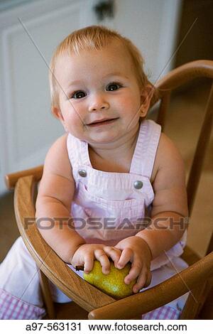 Happy blonde infant girl sitting in a high-chair while holding a green apple. View Large Photo Image Stock Image - Happy blonde infant girl sitting in a high-chair while holding a green apple.. Fotosearch