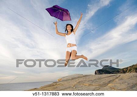 Happy brunette woman with umbrella jumping in a cliff by the sea in Ibiza View Large Photo Image Stock Image - Happy brunette woman with umbrella jumping in a cliff by the sea in Ibiza. Fotosearch