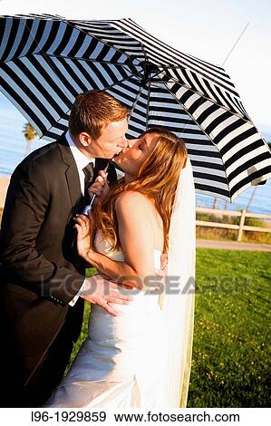 Stock Photo - happy couple after wedding ceremony, kissing and embracing under striped, black/white umbrella @ beach. Fotosearch
