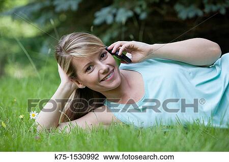 Stock Image - Happy woman lying down on lawn talking on cell phone. Fotosearch
