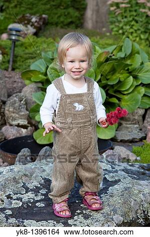 Picture - Happy Young Caucasian Kid Girl Holding Flowers and Standing on Stone in Garden. Fotosearch