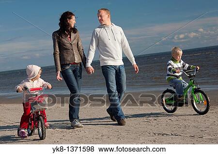 Stock Photo - Happy Young Family Holding Hands Walking with Kids on Bicycles by Sea, P? rnu Beach, Estonia. Fotosearch