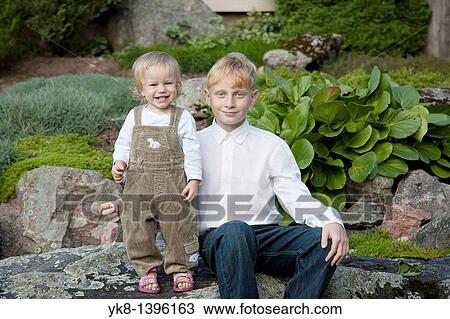 Stock Image - Happy Young Teenage Caucasian Boy with Kid Sister on Stone in Garden. Fotosearch