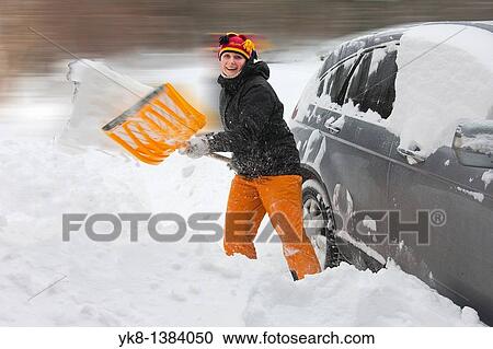 Stock Image - Happy Young Woman Digging Out a Car Using Snow Shovel. Fotosearch