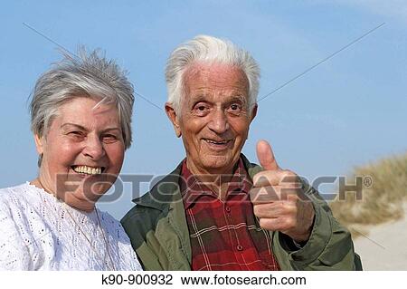 Stock Image - Head and shoulder photograph of elderly caucasian couple smiling and looking into the camera with blue sky and thumbs up gesture, Island of Amrum, Northfrisian, Schleswig_Holstein, Northern Germany, Europe. Fotosearch