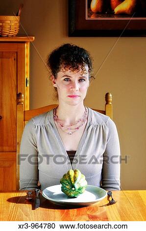 Stock Image - Head and shoulders portrait of a woman contemplating a whole squash on her dinner plate. Fotosearch