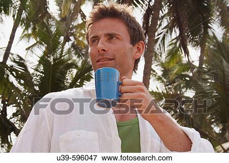 Stock Photo - Horizontal shot of a good-looking man holding a mug of coffee with palm-trees in the background, shot in India.. Fotosearch
