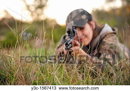Stock Image - Hunting woman lying on the grass is about to shoot a rifle firearm. Fotosearch