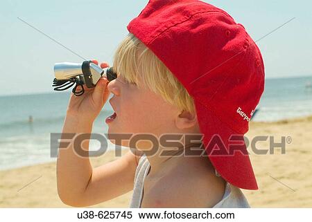 Stock Photography - In the sand along the Atlantic Ocean young boy age 3-4 sitting in the sand looking through a small telescope at the Atlantic ocean wearing a red hat. Virginia Beach, Virginia. USA. Fotosearch