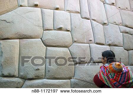 Stock Photo - Inca woman with child in traditional dress looking at Inca Roca's palace, now part of the Archbishop's Palace in the Hatun Rumiyoc street. The main feature is a twelve-angled stone in the middle of the wall. Cusco, Peru. Fotosearch