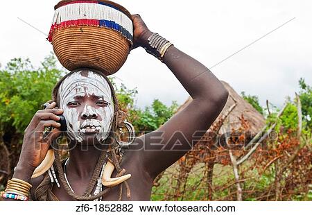 Stock Image - Jinka Ethiopia Africa village Lower Omo Valley Mago National Park wild tribe Mursi tribal woman with basket portrait on cell phone communicating with paint on face and traditional culture 15. Fotosearch