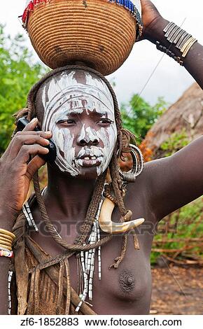 Stock Image - Jinka Ethiopia Africa village Lower Omo Valley Mago National Park wild tribe Mursi tribal woman with basket portrait on cell phone communicating with paint on face and traditional culture 15. Fotosearch