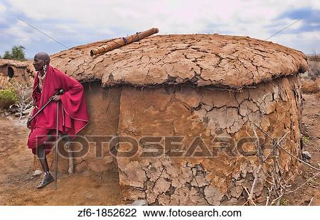 Stock Image - Kenya Africa Amboseli Maasai tribe village Masai man in red costume dress and beads in front of mud home in remote area of Amboseli National Park safari 1. Fotosearch