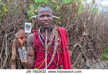 Stock Image - Kenya Africa Amboseli Maasai tribe village Masai man in red costume dress and beads holding Fuji Polaroid in remote area of Amboseli National Park safari 1. Fotosearch