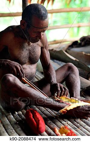 Picture - Kombai man sitting on the floor and preparing a breadfruit, Papua, Indonesia, Southeast Asia.. Fotosearch