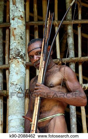 Stock Image - Kombai man standing in front of his wooden house with bow and arrows, Papua, Indonesia, Southeast Asia.. Fotosearch