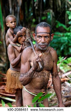Stock Image - Kombai man standing in the jungle with stone axe his wife with kid in the background, Papua, Indonesia, Southeast Asia.. Fotosearch