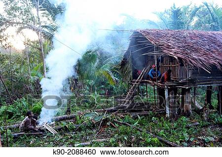 Stock Image - Kombai people sitting in front of a traditional house in the early morning, Papua, Indonesia, Southeast Asia.. Fotosearch