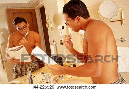 Stock Image - Latin man brushing his teeth while reading a magazine in a hotel room bathroom. Fotosearch