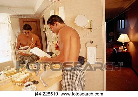 Stock Photo - Latin man brushing his teeth while reading a magazine in a hotel room bathroom. Fotosearch