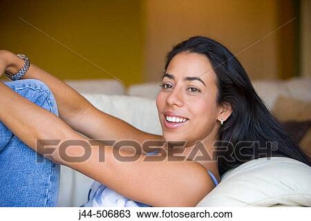 Stock Image - Latin woman sitting on couch smiling. Fotosearch